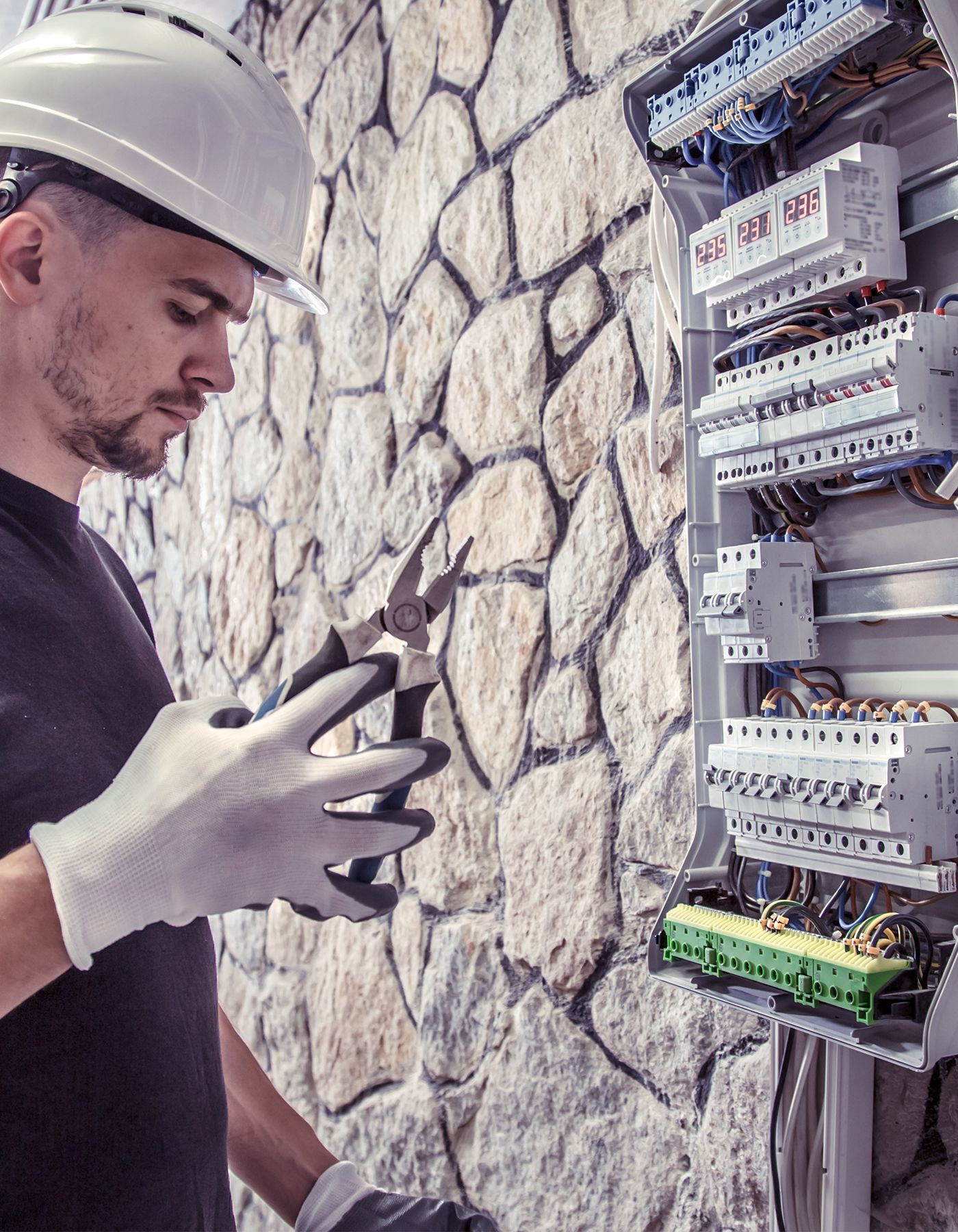 Elektriker i vit hjälm arbetar med kopplingsskåp vid stenvägg