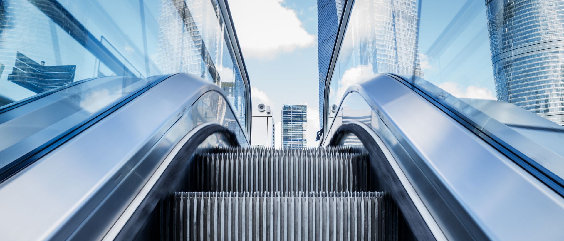 Rulltrappa sedd nedifrån med glasbyggnader mot blå himmel